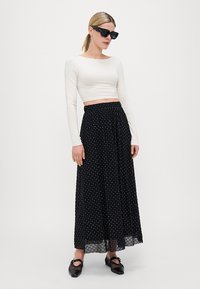 Svart maxi-skjørt med hvite prikker og gjennomsiktig overlag, kombinert med en tettsittende langermet hvit crop top. Modellen har på seg mørke solbriller og svarte sko.