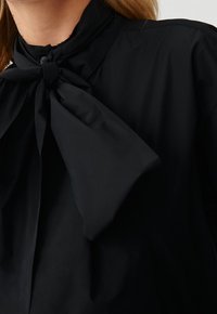 Schwarze Bluse mit hohem Kragen und einer großen Schleife am Hals, aus glattem Stoff gefertigt, zeigt klare Linien und ein minimalistisches Design.