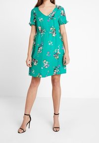 Kurzes türkisfarbenes Kleid mit V-Ausschnitt, Rüsche-Ärmel, blumigen Mustern in verschiedenen Farben und einer lockeren Passform, kombiniert mit schwarzen Riemchen-Heels.