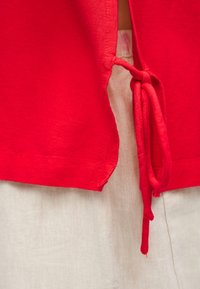 Ενδυμα από κόκκινο ύφασμα με πλαϊνή σχισμή δεμένη με κόμπο πάνω σε φόντο από ανοιχτό μπεζ υφή υφάσματος.