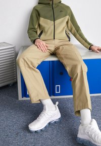 Persona con chaqueta Nike verde y beige, pantalones beige, calcetines blancos y zapatillas blancas sentada en un banco de taquilla azul en interiores.