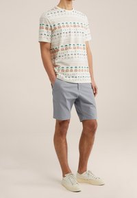 Koszulka krótki rękaw, jasny kolor, z wzorami przedstawiającymi palmy i geometryczne kształty, sparowana z szarymi szortami i jasnymi sneakerami.