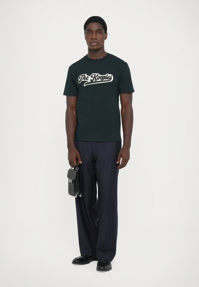 Melnas kokvilnas T-krekls ar baltu grafisku tekstu, apvienots ar plisētām tumši zilo svītraino biksēm un melniem ādas apaviem, turot nelielu melnu somiņu.