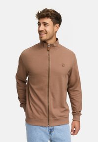 Hombre sonriendo, vistiendo una chaqueta marr�ón de cremallera con textura de canalé y jeans de mezclilla azul claro, de pie frente a un fondo blanco liso.