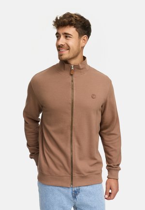 Hombre sonriendo, vistiendo una chaqueta marrón de cremallera con textura de canalé y jeans de mezclilla azul claro, de pie frente a un fondo blanco liso.
