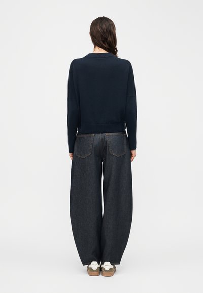 Personne portant un pull bleu marine, un jean denim foncé à jambes larges et des baskets blanches, debout de dos face à la caméra sur un fond uni.