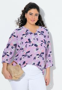 Una blusa floreale lavanda con maniche a tre quarti, caratterizzata da accenti blu navy e rosa, con un collo a V e bottoni, abbinata a una borsa intrecciata beige.