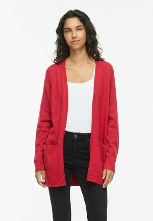 Femme aux cheveux bouclés foncés portant un cardigan rouge ouvert, un haut blanc et un jean noir, debout devant un fond blanc uni.