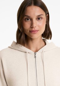 Giovane donna con capelli castani che indossa una felpa con zip beige e coulisse, fissando la telecamera su uno sfondo bianco.