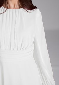 Robe blanche à manches longues avec un col froncé et des détails plissés, présentant une taille ajustée et une texture de tissu lisse et fluide.