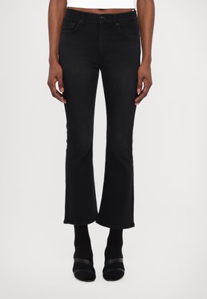 Schwarze, weit geschnittene Jeans mit hoher Taille und Vorderentaschen, die eine glatte Denim-Oberfläche und einen sauberen Stich entlang des Saums aufweisen. Kombiniert mit schwarzen Schuhen mit Knöchelriemen.