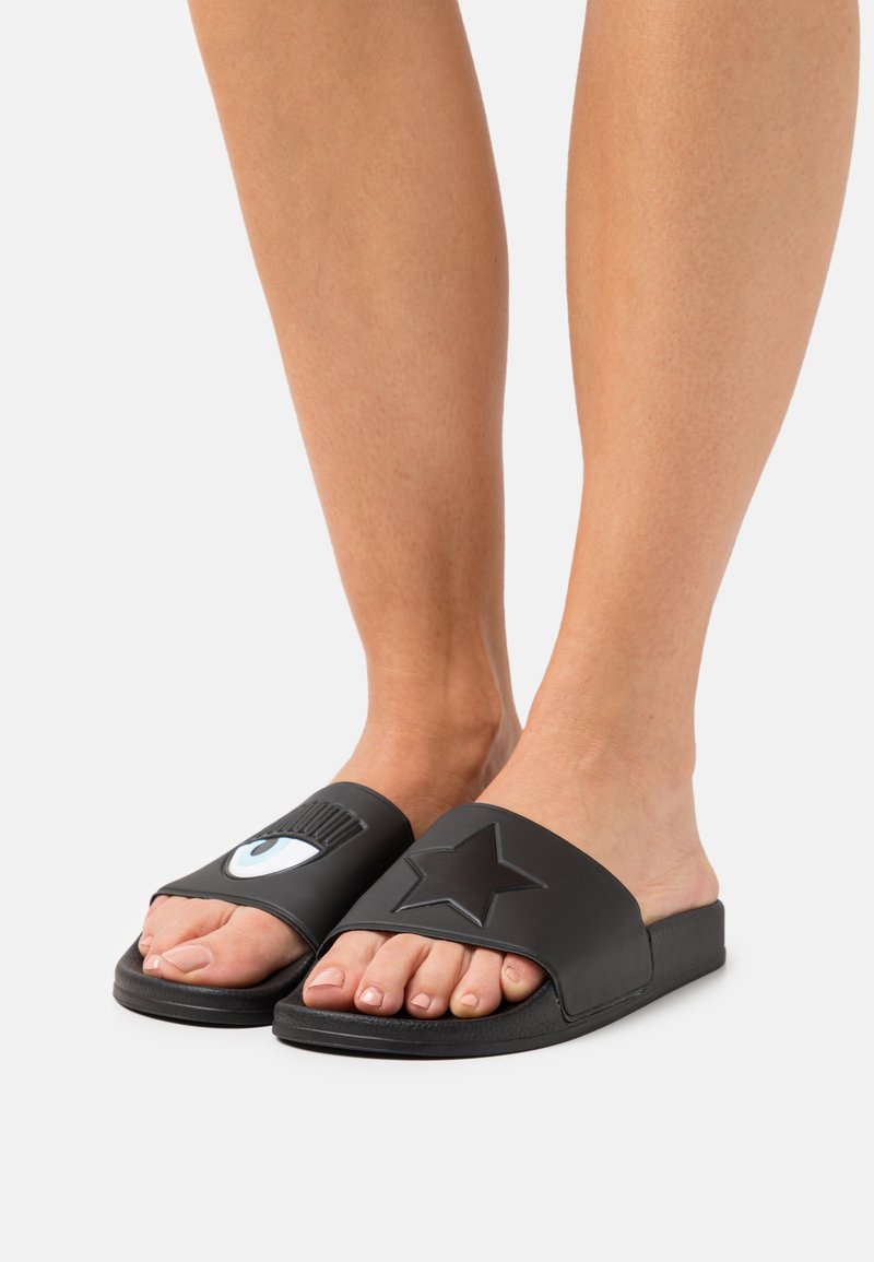 Schwarze Rutschsandalen mit einem breiten Riemen, der auf der einen Seite ein blaues Augenmotiv und auf der anderen Seite ein Sternenmuster zeigt. Strukturierte Gummisohle.