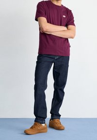 Camiseta de cuello redondo color burdeos, jeans oscuros con costuras en contraste y zapatillas de ante marrón. Ajuste casual y diseño sencillo.