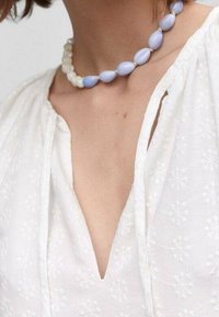 Witte katoenen blouse met bloemenborduursel, met een diepe V-hals. Draagt een korte ketting met afwisselend lichtblauwe en witte kralen.