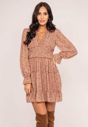 Femme aux longs cheveux foncés portant une robe marron à manches longues à motif floral et des bottes marron montant jusqu'aux genoux, debout avec une main sur la hanche.