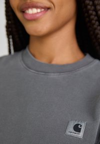 Grå bomullströja med rund halsringning, utrustad med en liten, fyrkantig Carhartt-logotyp på bröstet. Slät textur.