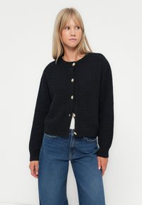 Svart strikket cardigan med teksturert overflate, rund hals og store gullknapper, kombinert med blå høyt liv jeans.
