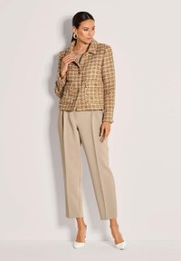 Vrouw in beige hoge taille broek, witte hakken en een gestructureerde bruine geruite knoopjasje met gouden oorringen, staande tegen een witte muur.