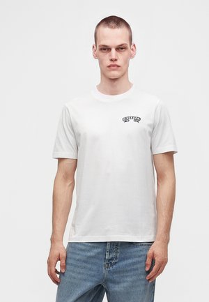 Joven con cabello corto que lleva una camiseta blanca de Diesel y vaqueros azules sobre un fondo claro y liso.