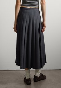 Una donna che indossa una gonna midi plissettata grigia, calze bianche arricciate, scarpe marroni stringate e un top grigio senza maniche, mentre si trova su un pavimento di cemento.
