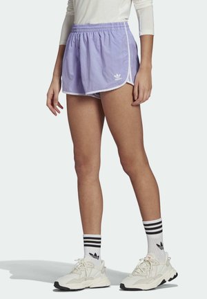 Persona vistiendo shorts deportivos lilas de Adidas, calcetines blancos de Adidas con rayas negras y zapatillas blancas, de pie frente a un fondo liso.