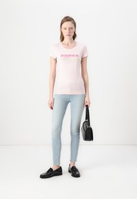 Camiseta ajustada rosa claro con el logo "GUESS LOS ÁNGELES" en colores degradados, combinada con jeans skinny azul claro y mocasines negros, sosteniendo un bolso negro.