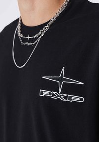 Camiseta de algodón negra con el logotipo bordado en blanco "PXP" y diseño de estrella en el lado izquierdo del pecho. Se llevan tres collares de cadena plateada en capas.