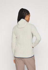 Eine Frau mit geflochtenem Haar, die einen hellgrauen, strukturierten Reißverschluss-Hoodie und beige Hosen trägt, steht mit einer Hand auf der Hüfte und schaut weg.