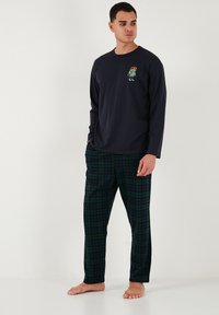 Kék hosszú ujjú crewneck póló hímzett karakterrel, zöld kockás pizsama nadrággal, amely puha pamutból készült. Mindkét ruhadarab lazán illeszkedik.