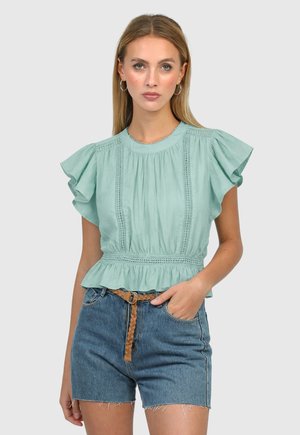 Femme portant un chemisier vert clair à manches courtes à volants avec des détails en dentelle et un short en jean taille haute avec une ceinture tressée marron.