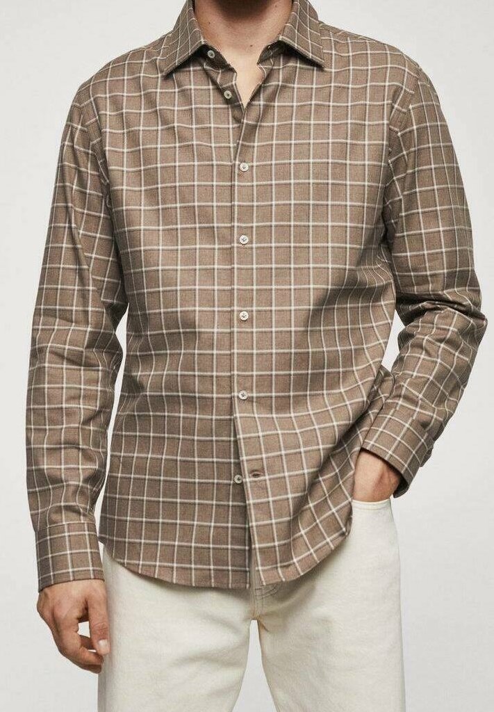 Homme portant une chemise à manches longues marron à carreaux avec des lignes blanches et un pantalon beige, une main dans la poche, debout devant un fond uni.