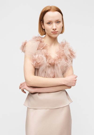 Jeune femme aux cheveux auburn courts portant une robe rose pâle ornée de détails en tulle floral transparent sur les épaules et le décolleté, les bras croisés.