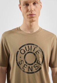 T-shirt en coton beige à manches courtes et col rond, arborant un grand graphisme circulaire noir avec le texte "QUITE SENSE" en police usée.
