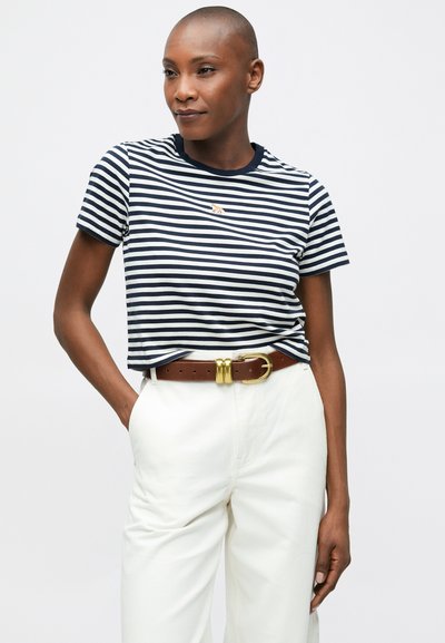 Modré a biele pruhované tričko s okrúhlym výstrihom, krátkymi rukávmi a malým vyšitým motívom na prsiach, skombinované s bielymi nohavicami s vysokým pásom a hnedým opaskom.