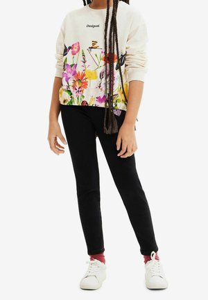 Witte sweatshrt met bloemmotief en kleurrijke prints, voorzien van een zwart logo, gecombineerd met zwarte broek en witte sneakers. Lang gevlochten haar zichtbaar.