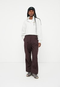 Pullover in pile grigio chiaro con colletto a mezza zip, abbinato a pantaloni cargo viola scuro con tasche laterali. Cappellino nero e sneaker grigie.