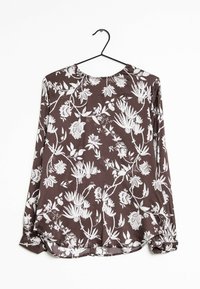 Langärmlige Bluse aus dunkelbraunem Stoff mit weißem Blumenmuster, Boot-Ausschnitt und gerafften Manschetten mit abgerundetem Saum. Hängt an einem schwarzen Haken.