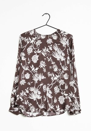 Blusa a maniche lunghe in tessuto marrone scuro con motivo floreale bianco, scollatura a barca e polsini arricciati con orlo arrotondato. Appesa a un gancio nero.