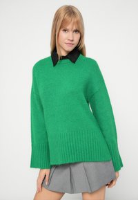 Νεαρή γυναίκα που φοράει ένα πράσινο πλεκτό πουλόβερ πάνω από ένα μαύρο πουκάμισο με γιακά και μια γκρι πλισέ φούστα, στέκεται μπροστά σε έναν απλό ανοιχτό φόντο.