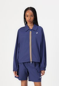 Navyblauwe lichte jas met een ritssluiting aan de voorkant, voorzien van gele en witte strepen, en losse mouwen. Gepaard met bijpassende navy shorts.