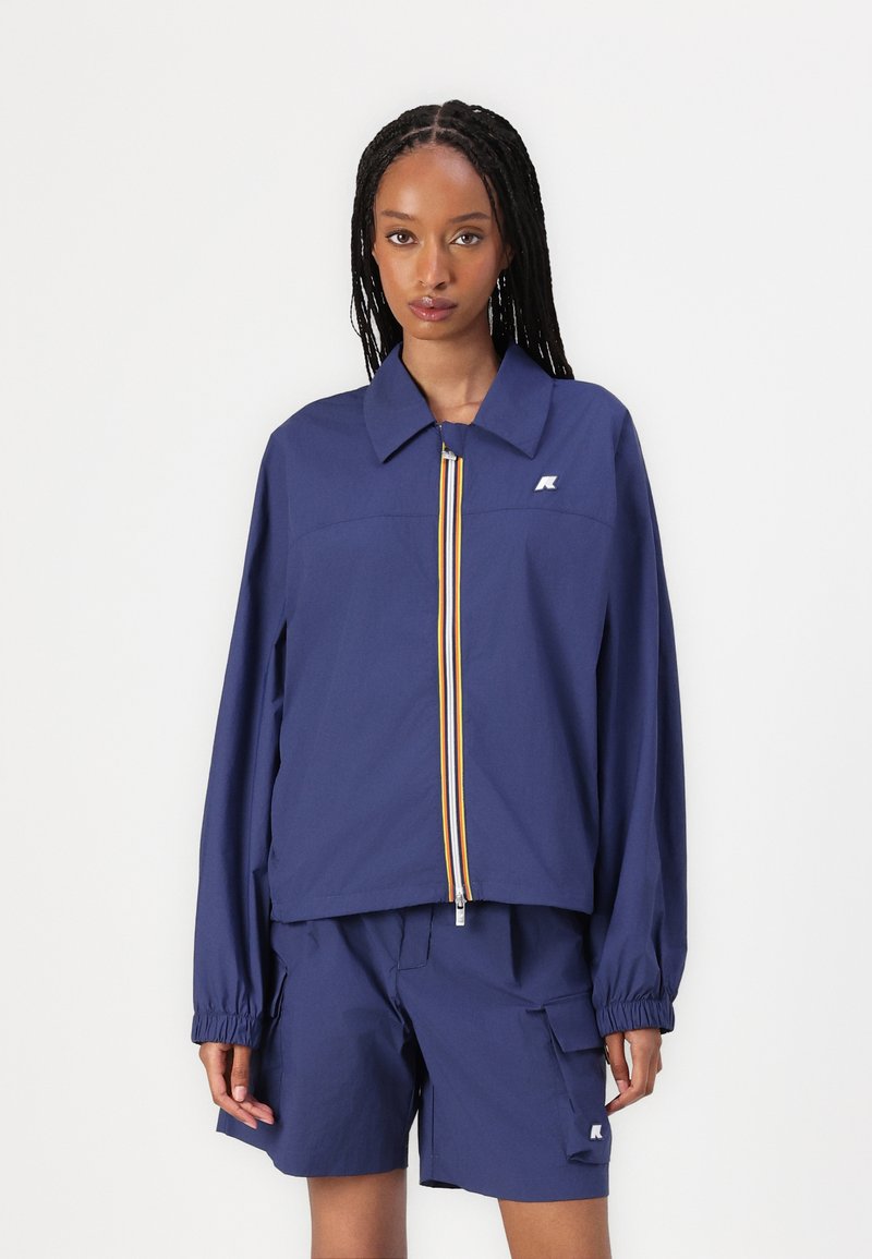 Navyblauwe lichte jas met een ritssluiting aan de voorkant, voorzien van gele en witte strepen, en losse mouwen. Gepaard met bijpassende navy shorts.