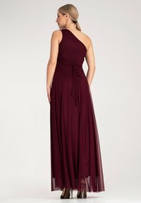 Burgundowa maxi sukienka na jedno ramię o zwiewnej sylwetce, z detalem związanym w talii i przezroczystymi akcentami. Gładka faktura i elegancki design.