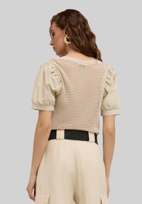 Top a maniche corte con un design in rete beige e maniche a sbuffo. Indossato sopra pantaloni a vita alta con una cintura nera texturizzata.