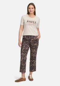 T-shirt beige clair à manches courtes avec un design de texte "JOYFUL" ; pantalon court à motifs marron et marine. Modèle portant des baskets marron.