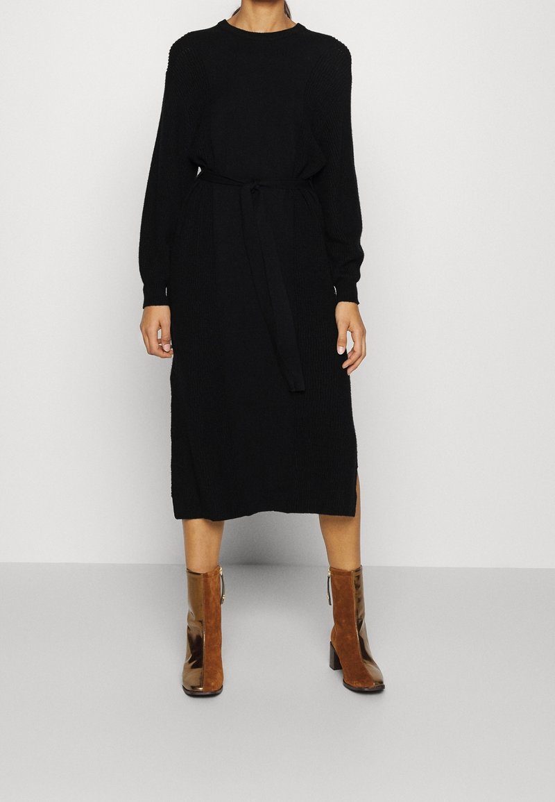 Femme en robe noire en maille à manches longues avec une ceinture à la taille et des bottines marron, debout devant un fond clair uni.