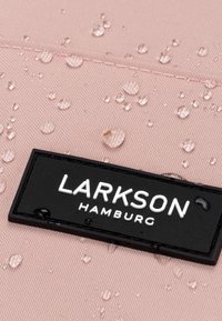 Ružová tkanina s vodnými kvapkami a čiernou obdĺžnikovou štítkou s nápisom "LARKSON HAMBURG" v bielej farbe. Viditeľný detail textúrovaného povrchu.