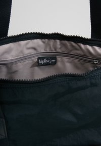 Sac Kipling noir avec zip ouvert montrant une doublure en tissu beige et une poche intérieure zippée avec une étiquette logo Kipling.