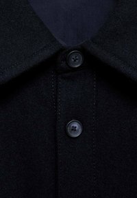 Donker wollen shirt met een puntige kraag en twee zwarte knopen. De stof heeft een gladde textuur en toont subtiele stiksels.