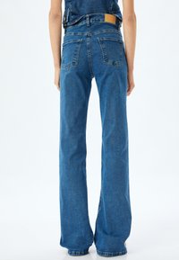 Vaqueros de tiro alto y pierna ancha en denim azul medio. Cuenta con bolsillos traseros y un parche de cuero marrón en la cintura.