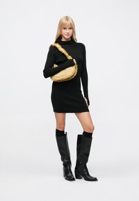 Rochie neagră cu mâneci lungi și aspect reiat, geantă galbenă cu franjuri pe umăr, cizme negre până la genunchi cu toc gros, pozând pe un fundal alb simplu.